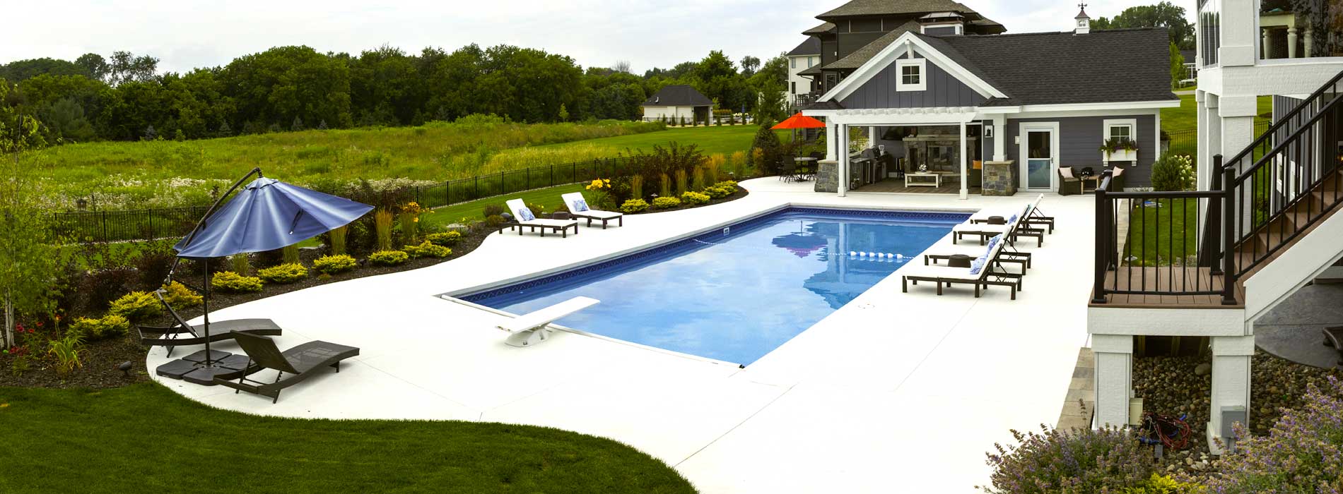 Panoramic view of a residential inground pool with white patio pavers, pool house, and landscaped yard in the Twin Cities, MN